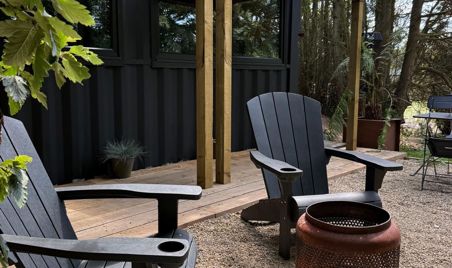 The Bolt Hole exterior at Coast and Castles — dark timber cladding, covered wooden porch, Adirondack chairs and gravel seating area